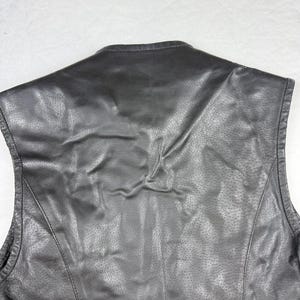 May include: Black leather vest with a textured surface and a subtle sheen. The vest features a classic design with a tailored fit, visible stitching, and a rounded neckline. The back of the vest is shown in the image.