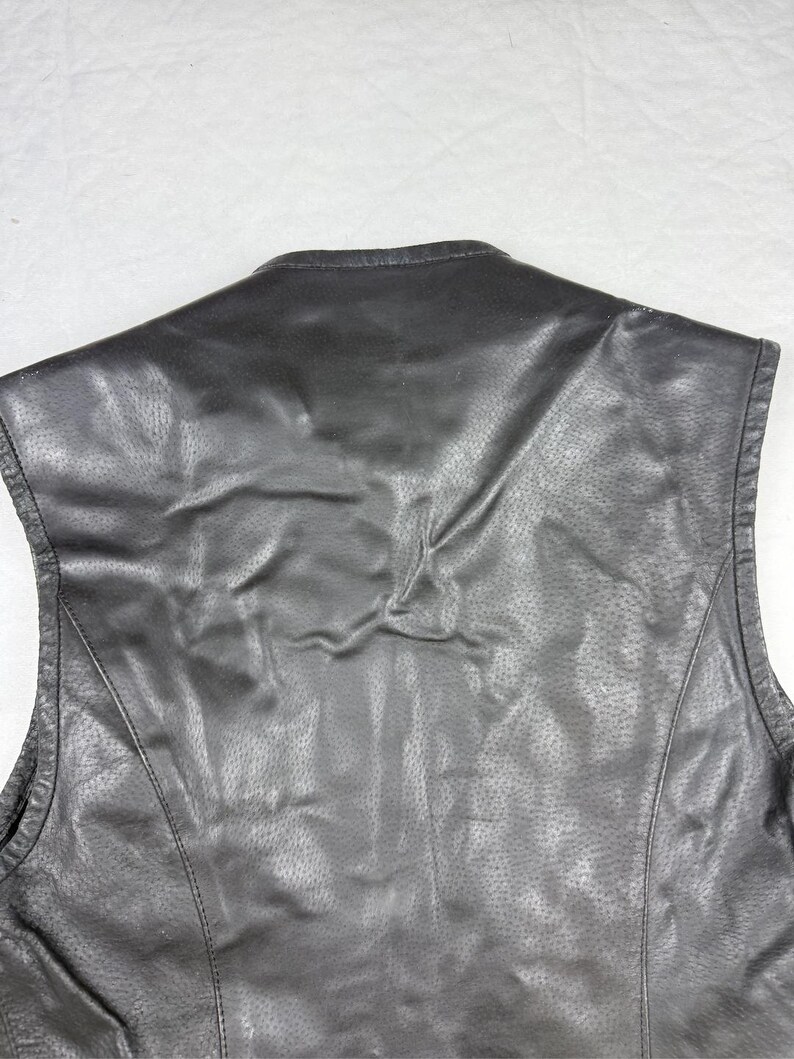 May include: Black leather vest with a textured surface and a subtle sheen. The vest features a classic design with a tailored fit, visible stitching, and a rounded neckline. The back of the vest is shown in the image.