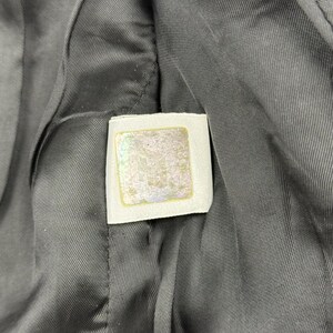 May include: Close-up of a gray fabric with a white label. The label has a square shape with a gold border and a faded design in the center. The fabric has a textured appearance.