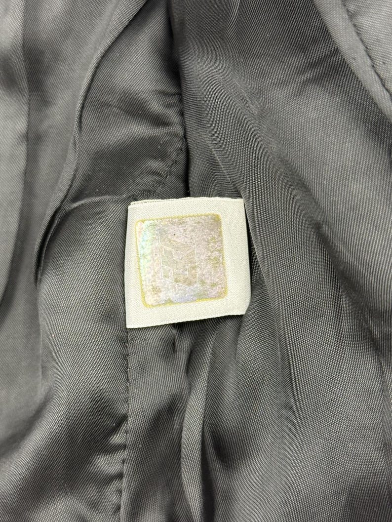 May include: Close-up of a gray fabric with a white label. The label has a square shape with a gold border and a faded design in the center. The fabric has a textured appearance.
