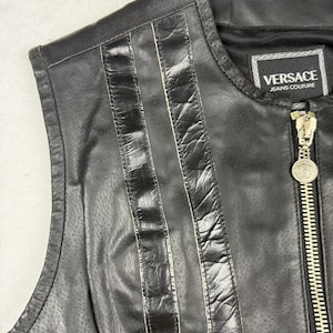 May include: Black leather vest with vertical stitched accents and a full zipper. The vest features the "Versace Jeans Couture" label. The zipper pull has a decorative medallion. The vest is sleeveless.