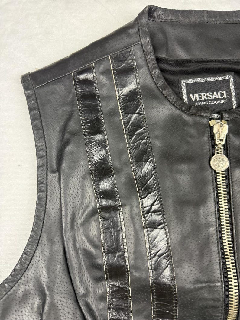May include: Black leather vest with vertical stitched accents and a full zipper. The vest features the "Versace Jeans Couture" label. The zipper pull has a decorative medallion. The vest is sleeveless.