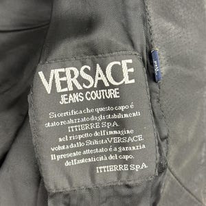 May include: Close-up of a black fabric label with the white text "VERSACE JEANS COUTURE." Below the logo is Italian text. A small blue tag with white text is visible on the right side. The label is sewn into a black garment.