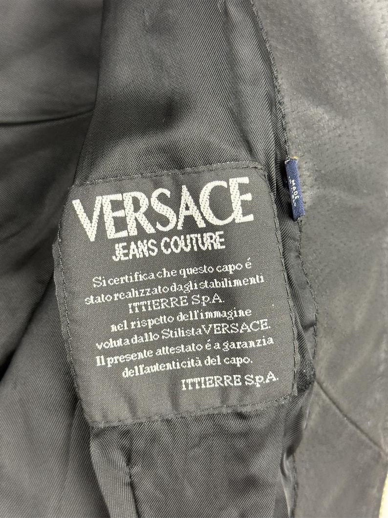 May include: Close-up of a black fabric label with the white text "VERSACE JEANS COUTURE." Below the logo is Italian text. A small blue tag with white text is visible on the right side. The label is sewn into a black garment.