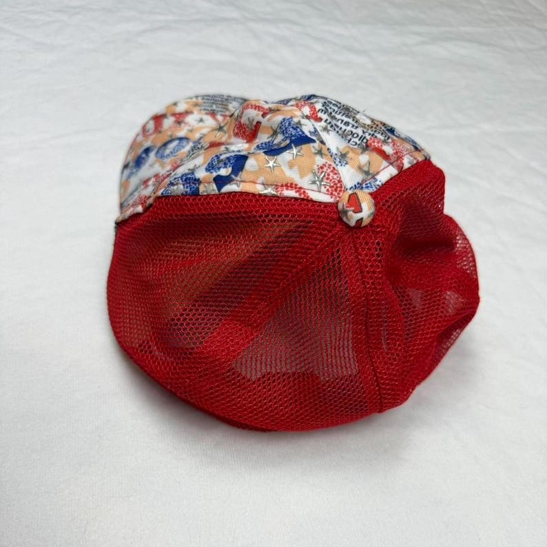 May include: A red mesh trucker hat with a patterned top featuring stars, text, and other patriotic designs in red, white, and blue. The hat has a curved brim and a button detail.