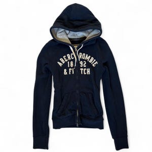 May include: A navy blue zip-up hoodie featuring a light grey interior hood lining and white drawstring cords. The front displays the white embroidered text Abercrombie 1892 & Fitch. It includes two front pockets and long sleeves with ribbed cuffs.
