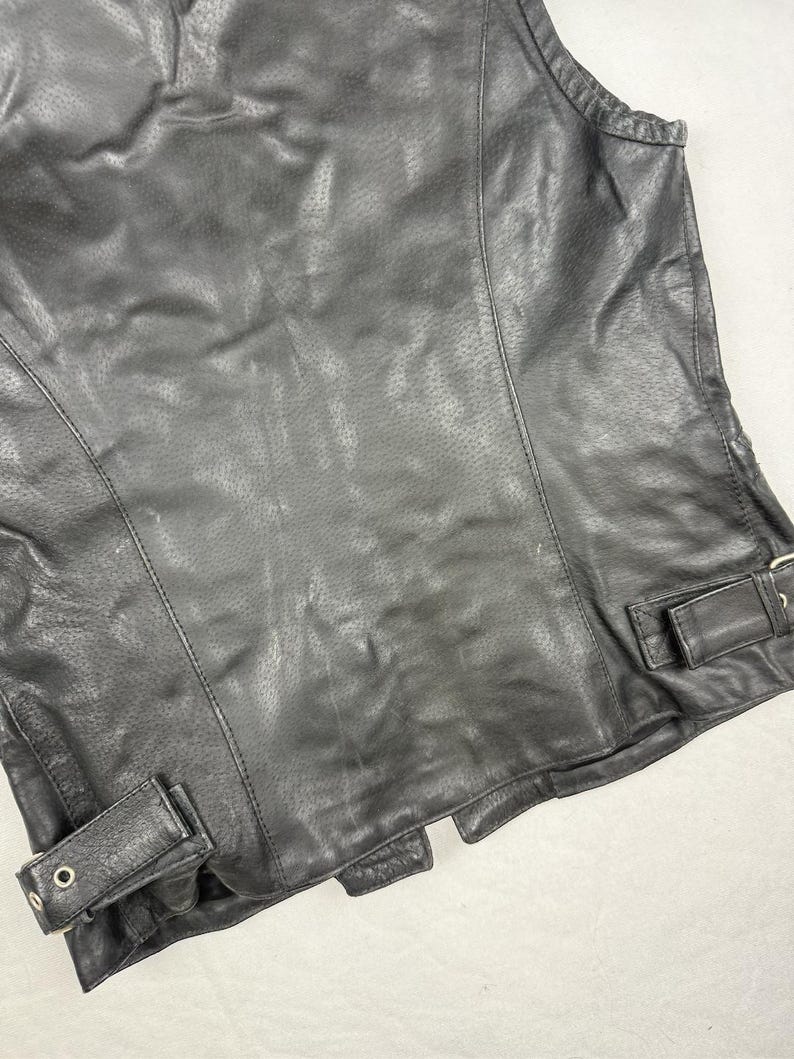 May include: Black leather vest with a perforated design. The vest features a classic biker style with a tailored fit, visible stitching, and adjustable side straps with metal buckles. A timeless piece for a rugged look.
