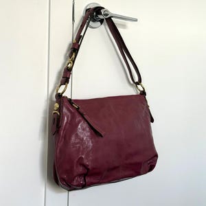 May include: A burgundy leather shoulder bag with a long strap and gold-colored hardware. The bag is hanging from a silver door handle against a white door. The bag has a rectangular shape and a soft, slouchy appearance.