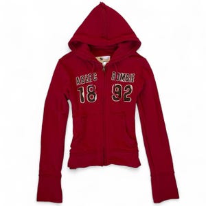 May include: A deep red zip-up hoodie featuring long sleeves and a hood. The front displays the text ABERCROMBIE 18 92 in white and dark brown lettering. The garment includes two front pockets and a central zipper closure extending from the hem to the neck.