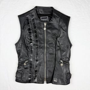 May include: Black leather vest with a zip-up front, featuring a round collar and multiple pockets. The vest has a decorative panel on the left side and a Versace logo patch near the collar. The vest is sleeveless.