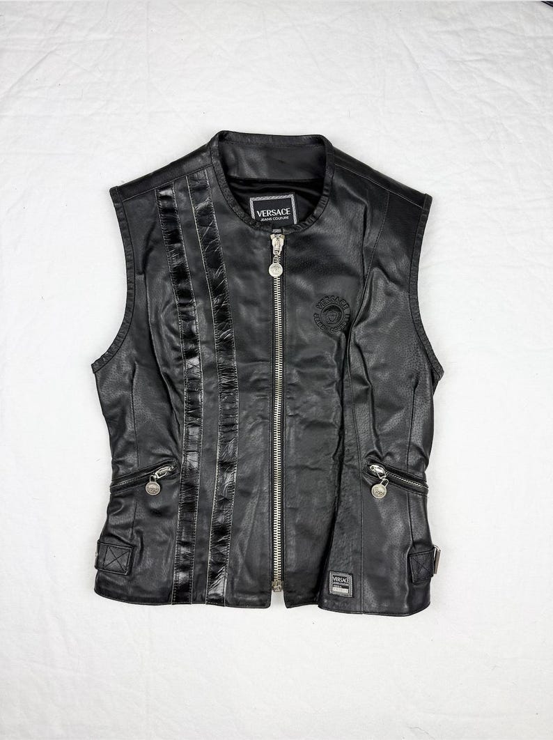 May include: Black leather vest with a zip-up front, featuring a round collar and multiple pockets. The vest has a decorative panel on the left side and a Versace logo patch near the collar. The vest is sleeveless.