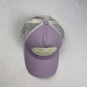 May include: A lavender baseball cap with a mesh back and a curved peak. The front of the cap features a patch with the text "Von Dutch" in a yellow and white design. The cap has a button on top.