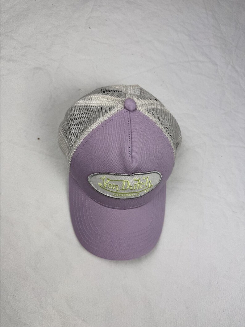 May include: A lavender baseball cap with a mesh back and a curved peak. The front of the cap features a patch with the text "Von Dutch" in a yellow and white design. The cap has a button on top.