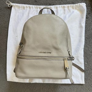 May include: A light gray leather backpack with silver-toned hardware, including zippers and a logo that reads "MICHAEL KORS". The backpack has a top handle and adjustable straps. It is sitting on top of a white drawstring bag.