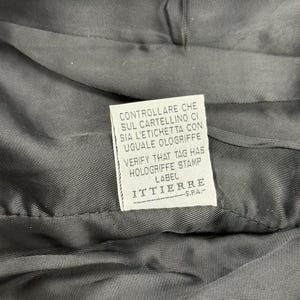 May include: Close-up of a dark gray fabric with a white label. The label has text in Italian and English, including "VERIFY THAT TAG HAS HOLOGRIFFE STAMP LABEL" and the brand name "ITTIERRE S.P.A."