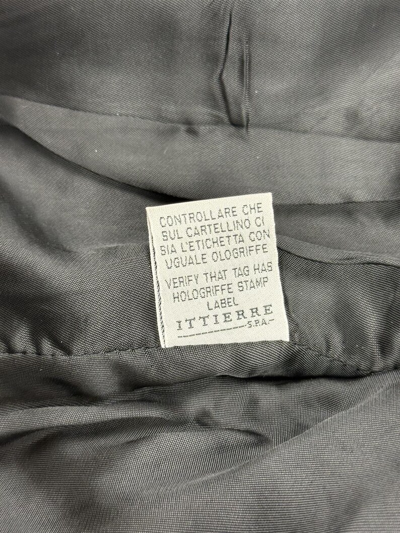 May include: Close-up of a dark gray fabric with a white label. The label has text in Italian and English, including "VERIFY THAT TAG HAS HOLOGRIFFE STAMP LABEL" and the brand name "ITTIERRE S.P.A."