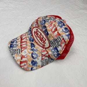 May include: A Von Dutch baseball cap with a red mesh back and a curved brim. The cap features a repeating pattern of stars, text, and shapes in red, blue, and tan. The Von Dutch logo is on the front.