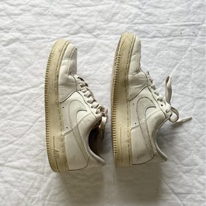 Pode incluir: Par de ténis Air Force 1 em branco sujo. Os sapatos têm um design clássico de cano baixo, com o icónico logótipo da Nike nas laterais. As solas e entressolas são de uma cor branco sujo ligeiramente envelhecida, e os atacadores são a condizer.