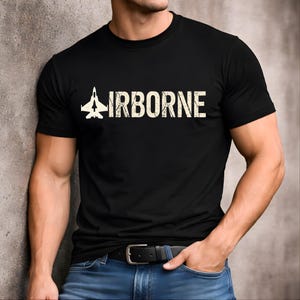 Puede incluir: Una camiseta negra de manga corta con un gráfico de la silueta de un avión de combate junto a la palabra AIRBORNE en una fuente desgastada de color blanco roto. El diseño está centrado en el pecho de la prenda, que tiene cuello redondo.