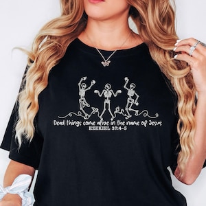 May include: A black t-shirt featuring a white line art graphic of three dancing skeletons. Below the skeletons, the text reads: Dead things come alive in the name of Jesus EZEKIEL 37:4-5. The design is centered on the front of the garment.