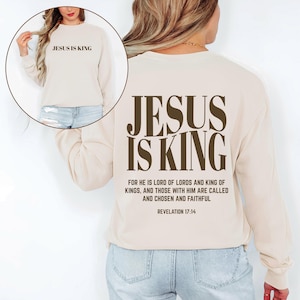 May include: Cream-colored sweatshirt with the text "JESUS IS KING" in brown on the front and back. The back also features a Bible verse. The sweatshirt is a long-sleeved, crew neck style.