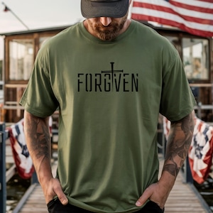 May include: Olive green t-shirt with the word "FORGIVEN" printed in black, featuring a cross design. The shirt has short sleeves and a crew neck. The background includes an American flag and a wooden structure.