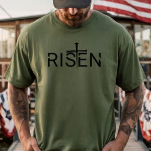 May include: Olive green t-shirt with the word "RISEN" in black, featuring a cross and nail design. The shirt is a short-sleeved crew neck style.