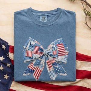 Op de afbeelding: Een blauw t-shirt met een groot strikdesign met de Amerikaanse vlag en het Vrijheidsbeeld. De strik is gemaakt van verschillende stofpatronen, waaronder sterren, strepen en ruitjes. Het shirt heeft een ronde hals en een label met de tekst "Comfort Colors".