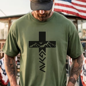 May include: An olive green short-sleeve t-shirt featuring a black graphic print of a cross. The cross contains a mountain range silhouette and a small bird, with the text G > ^ v below the mountains, centered on the chest of the garment.