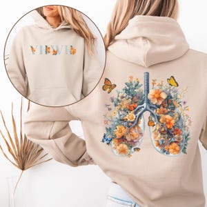 May include: Beige hoodie with a front pocket and drawstring hood. The front features the text "YHWH" in light blue with orange floral accents. The back displays a detailed illustration of lungs filled with orange flowers, greenery, and butterflies.