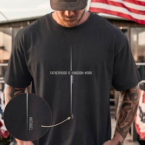 May include: Dark grey t-shirt with the text "FATHERHOOD IS KINGDOM WORK" in white. A vertical white line runs down the centre, with "MICHAEL" in a circle at the bottom. The shirt is worn by a person.