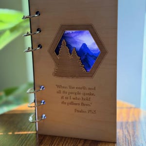 May include: Wooden notebook with a laser-cut mountain and tree design. The notebook features a hexagonal window revealing a blue and purple mountain scene. The text "When the earth and all its people quake, it is I who hold its pillars firm." is inscribed.