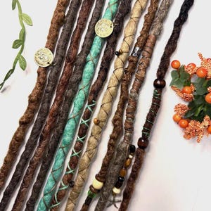 May include: A selection of handmade dreadlocks in shades of brown, black, and teal. Some dreads are decorated with beads, leather wraps, and metal charms. The dreadlocks are arranged on a white surface, highlighting their texture and design.