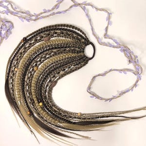 May include: A collection of brown, blonde, and black synthetic dreadlocks, secured with a black elastic band. Wooden beads are interspersed throughout the dreads. A decorative garland with purple leaves and twine is arranged around the dreadlocks.