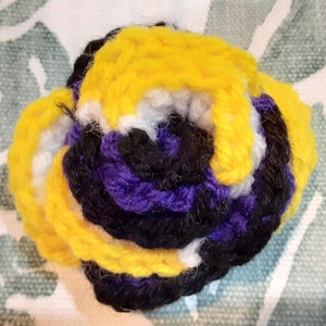 Op de afbeelding: Een handgemaakte gehaakte bloem in de kleuren van de non-binaire pride-vlag: geel, wit, paars en zwart. De bloem is strak gewikkeld, waarbij elke kleur concentrische cirkels vormt. Het garen lijkt zacht en getextureerd.