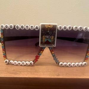 May include: Oversized sunglasses with a gradient lens, embellished with colorful rhinestones and beaded text that reads "YOU GOTTA TRY TO SEE A LITTLE FURTHER." A tarot card is centered on the bridge.