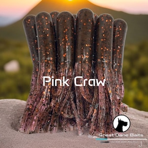 May include: A close-up of a bundle of pink crawfish-shaped fishing lures with brown and orange glitter. The lures are arranged in a circle and the text "Pink Craw" is written in white letters on a black background.