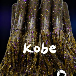 May include: A cluster of soft plastic fishing lures. The lures are dark brown with purple and yellow glitter. The word "Kobe" is written in white across the image. The Great Dane Baits logo is in the bottom right corner.