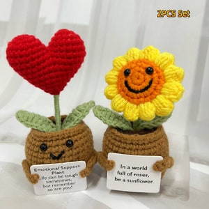 May include: Two crocheted plant pots, one with a red heart and the other with a yellow sunflower. Both pots have a smiling face and are holding signs with inspirational messages. The sign on the heart pot reads "Emotional Support Plant Life can be tough sometimes, but remember so are you!" The sign on the sunflower pot reads "In a world full of roses, be a sunflower."