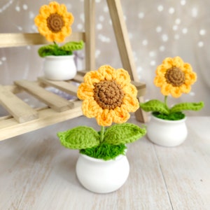 Può includere: Girasoli all'uncinetto in vasi di ceramica bianca. I girasoli hanno petali gialli e centri marroni. Foglie verdi e materiale simile al muschio circondano gli steli. I fiori sono esposti su una mensola di legno.