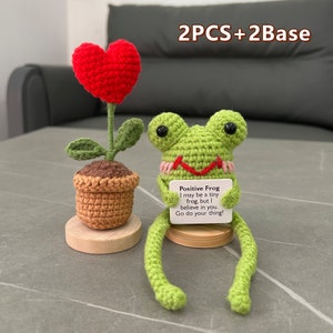 May include: Two crocheted amigurumi crafts: a green frog holding a sign that reads "Positive Frog I may be a tiny frog, but I believe in you. Go do your thing!" and a potted plant with a red heart-shaped flower.