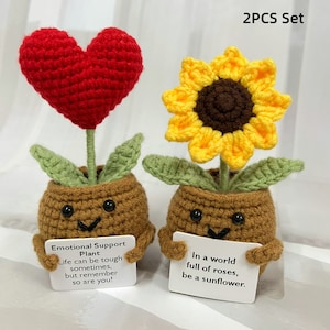 May include: Two crocheted plant pots with cute faces. One pot has a red heart flower and the other has a yellow sunflower. The pots are holding signs with the text "Emotional Support Plant Life can be tough sometimes, but remember so are you!" and "In a world full of roses, be a sunflower."