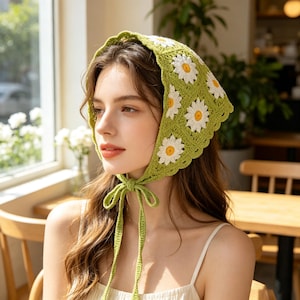 May include: A crochet headscarf featuring a green base with white and yellow daisy flower motifs. The headscarf is triangular in shape with a scalloped edge and includes two long green crochet ties that fasten under the chin to secure the piece.