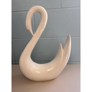 May include: A white ceramic swan ornament. The swan has a long, curved neck and a graceful, flowing form. The ornament is a decorative item, suitable for home decor.