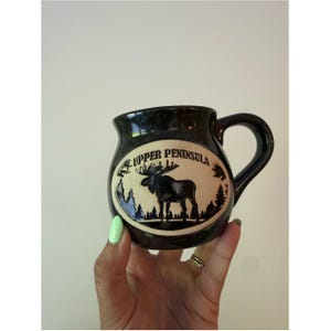 May include: A dark brown ceramic mug with a rounded shape and a side handle. The front features an oval cream-colored inset with a black silhouette of a moose standing in a forest, accompanied by the text UPPER PENINSULA printed in black.