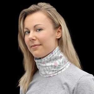 May include: A white neck warmer featuring a floral design with pink flowers and green leaves. The neck warmer is worn around the neck and is made of a soft material. The background is black.
