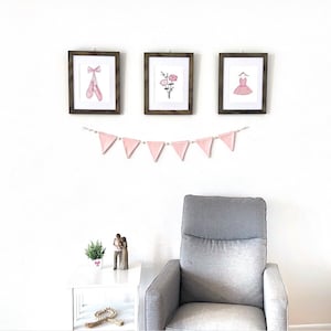 May include: Three framed prints with pink and white illustrations of ballet shoes, flowers, and a dress hang on a white wall. A pink and white bunting hangs below the frames. A gray armchair sits in front of the wall.