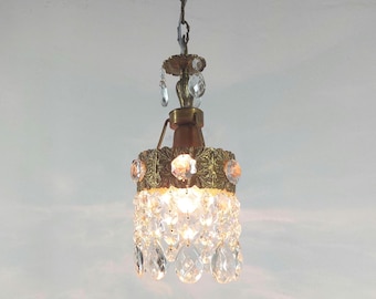 May include: A gold chandelier with crystal drops hangs from a chain. The chandelier has a decorative, ornate design and is lit with a warm, white light.