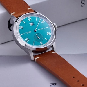 May include: A silver wristwatch with a teal face and brown leather band. The watch face features white numerals and hands, with a small subdial and date window. The band is stitched with white thread.