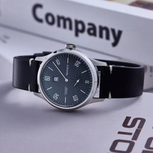 May include: A silver-toned wristwatch with a black leather band. The watch face is dark green with white numerals and hands. The watch has a date display and the text "MuFORTIC" is visible. The word "Company" is in the background.